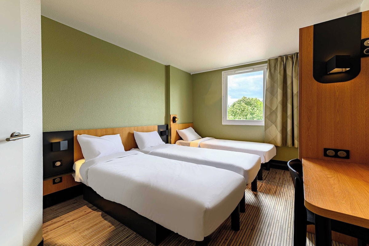 A hotel room with two beds, a wooden desk, a window with curtains, and a view of greenery outside.