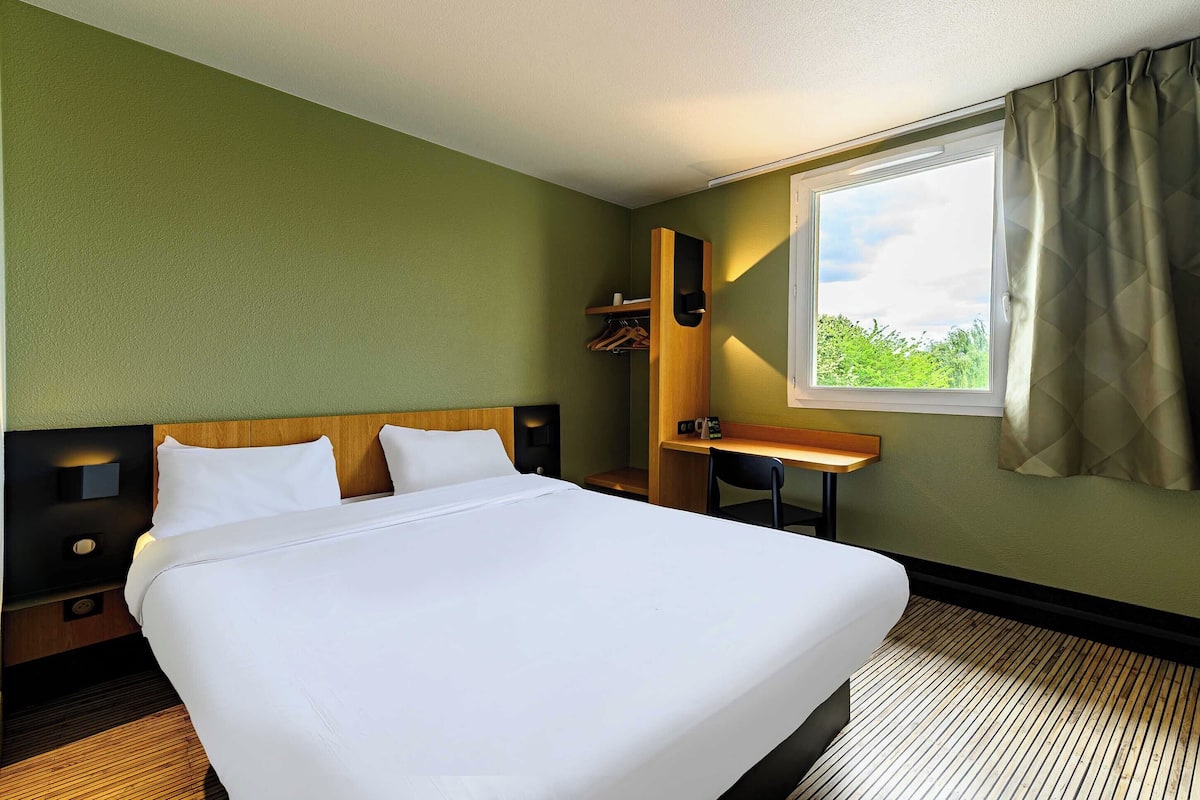 A hotel room with a large bed, a desk, a chair, a wardrobe, and a window with curtains.