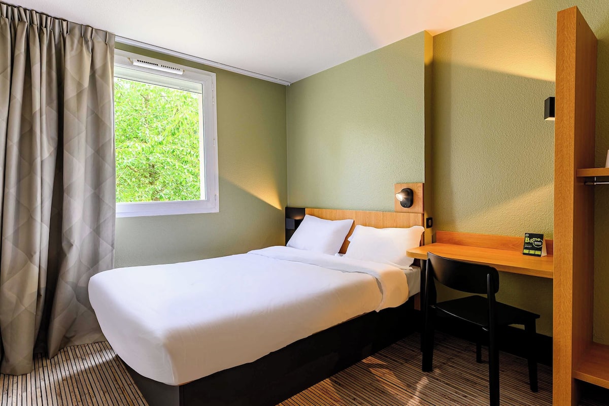 A hotel room with a bed, a desk, a chair, and a window with curtains.
