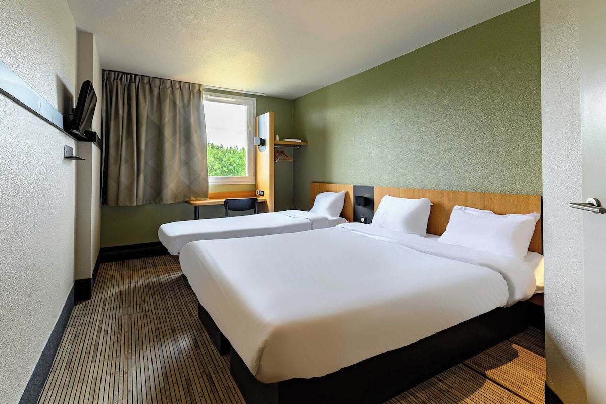 A hotel room with two beds, a desk, a chair, a window with curtains, and a view of greenery outside.