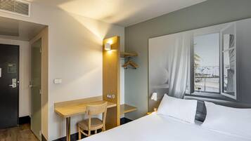 A hotel room with a bed, a desk, a chair, a mirror, a window, and a door.