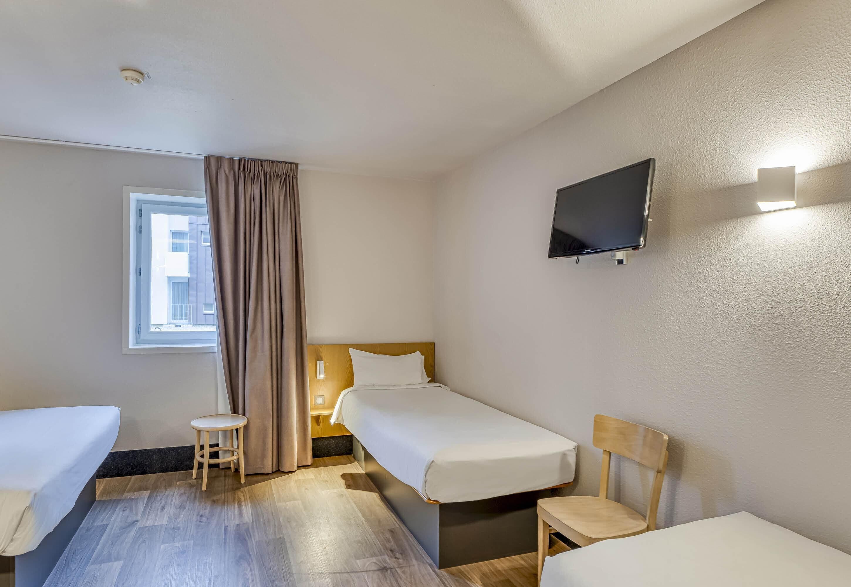 Habitación de hotel con dos camas individuales, una mesita, una silla, un televisor montado en la pared y una ventana con cortinas.