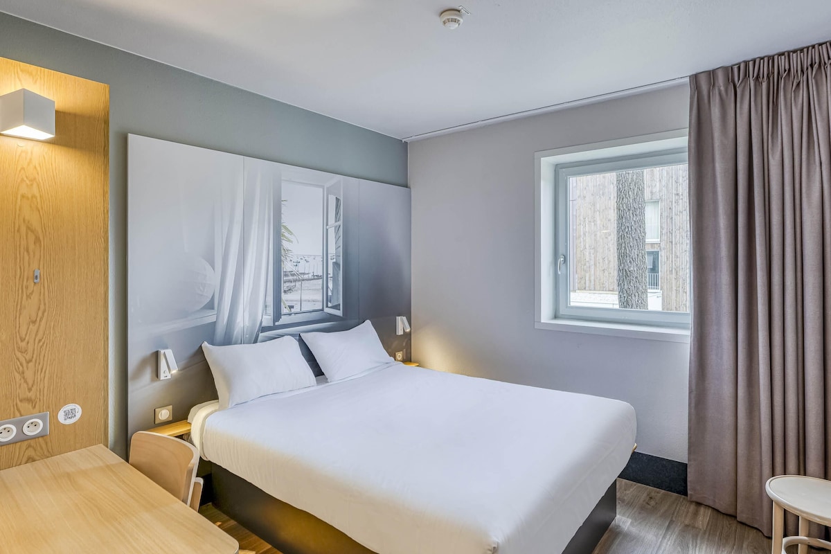 Hotelkamer met een bed, nachtkastje, lamp, raam met gordijnen en uitzicht.