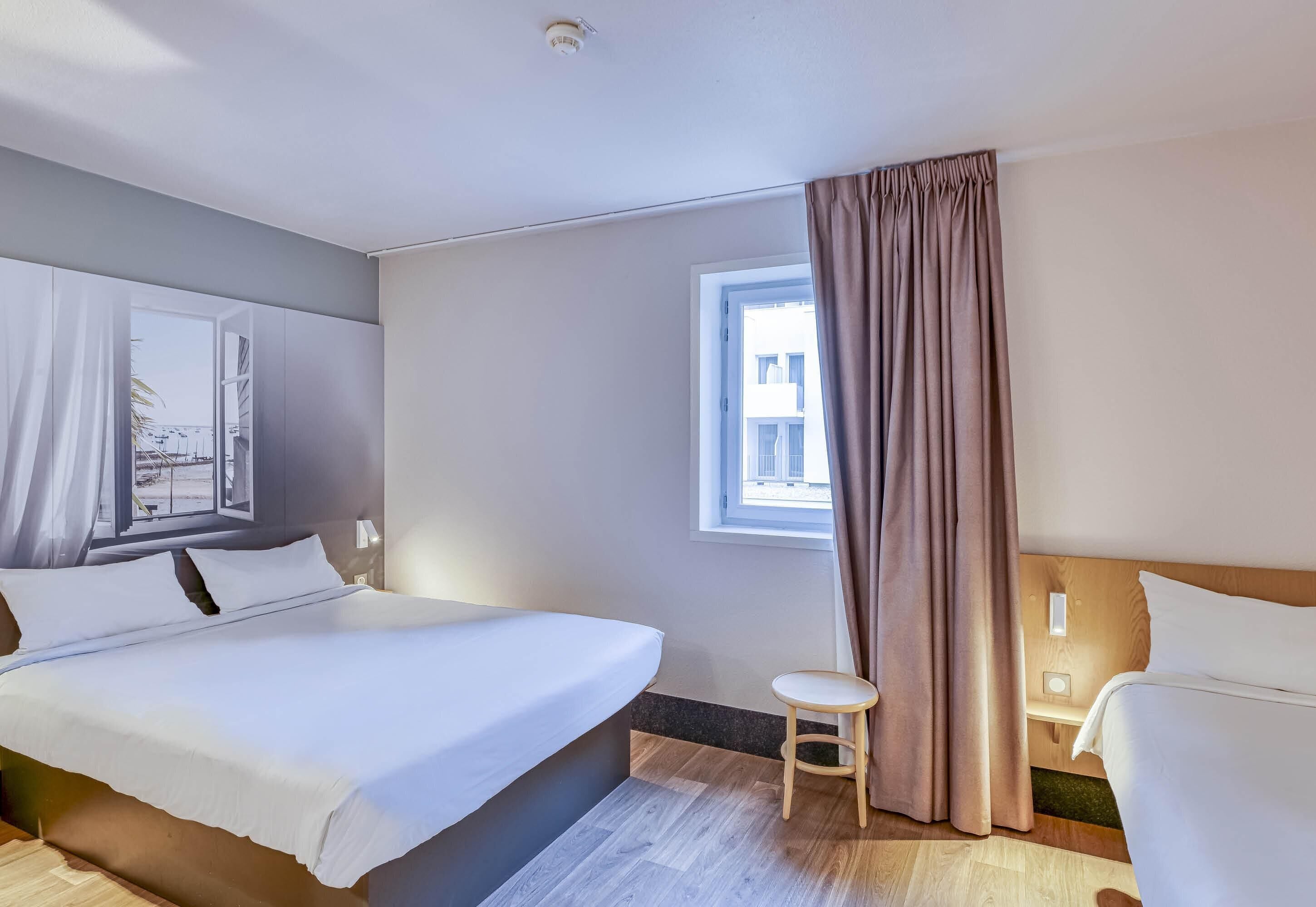 A hotel room with a bed, a small stool, a window with curtains, and a view of the outside.