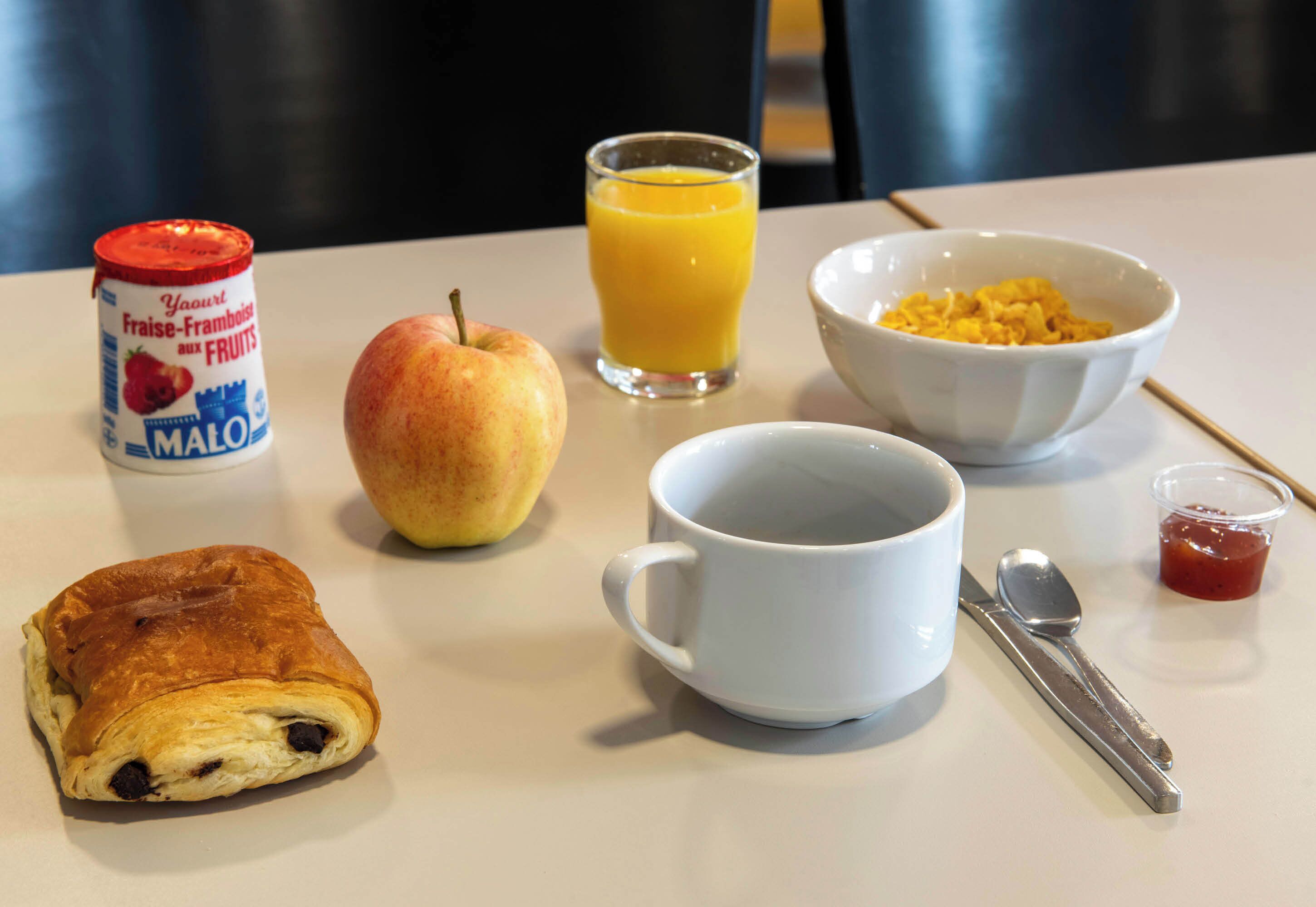 Una mesa de desayuno con un croissant, una manzana, un vaso de jugo de naranja, un tazón de cereal, una taza, una cuchara y un frasco de mermelada.
