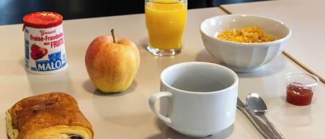 A breakfast table with a croissant, an apple, a glass of orange juice, a bowl of cereal, a mug, a spoon, and a container of jam.