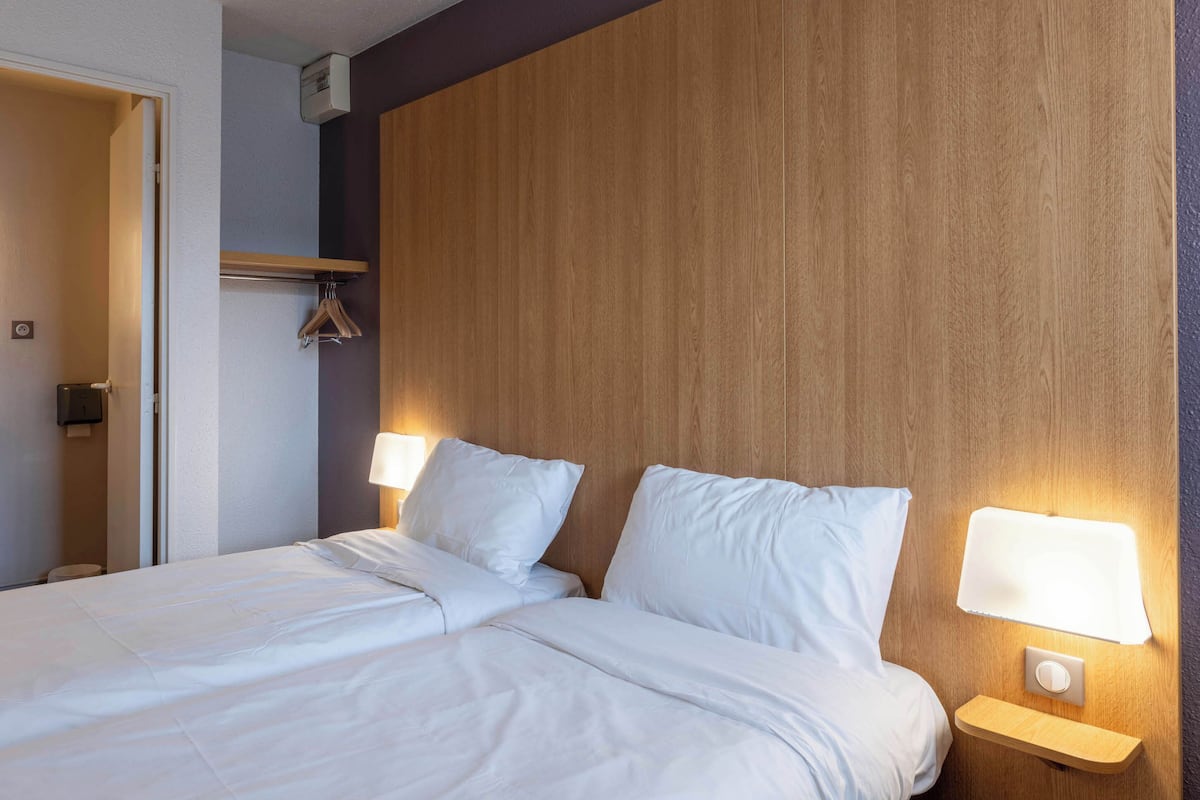 Hotellihuone, jossa on puinen sängynpääty, kaksi yöpöytävalaisinta, valkoisilla lakanoiden päällä varustettu sänky ja naulakko.