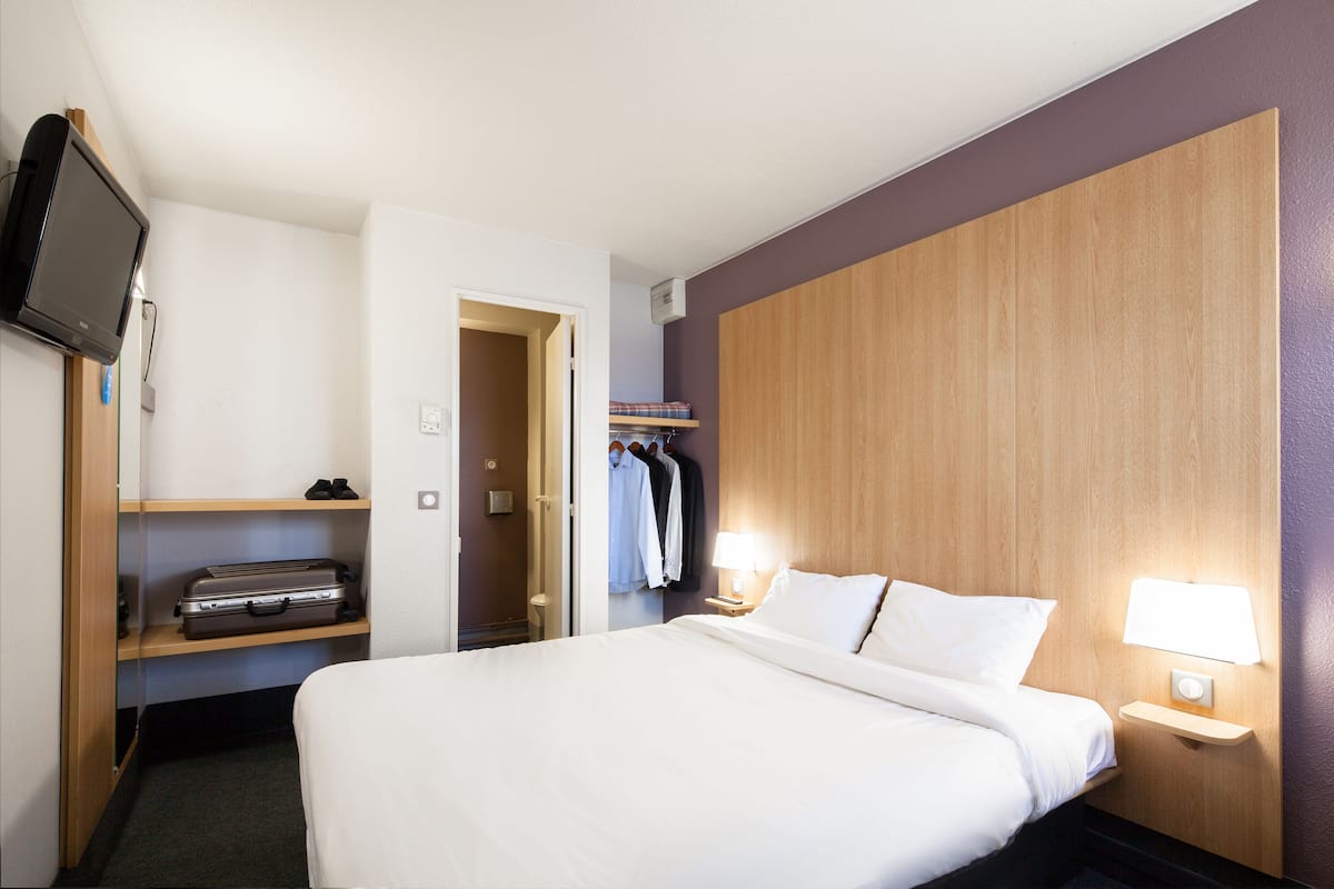 Hotellihuone, jossa on sänky, televisio, vaatekaappi ja yöpöytä, jossa on lamppu.