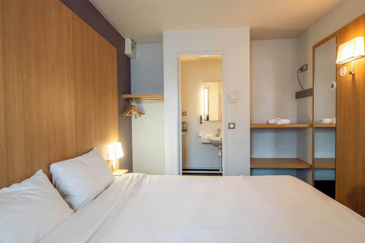 Hotellihuone, jossa on sänky, puinen pääty, yöpöytälamppu, peili, vaatekaappi ja kylpyhuone, joka näkyy avoimesta ovesta.