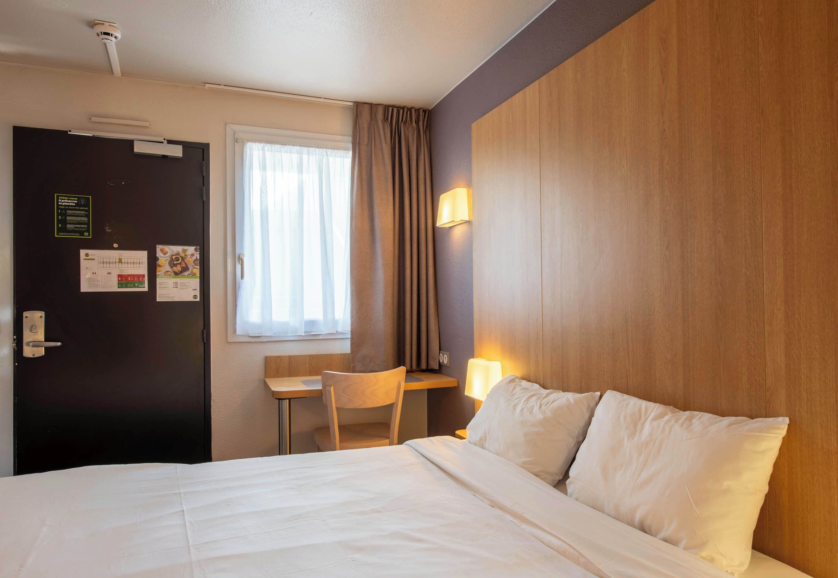 A hotel room with a bed, a desk, a chair, a window with curtains, and a wall-mounted lamp.