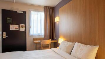 A hotel room with a bed, a desk, a chair, a window with curtains, and a wall-mounted lamp.