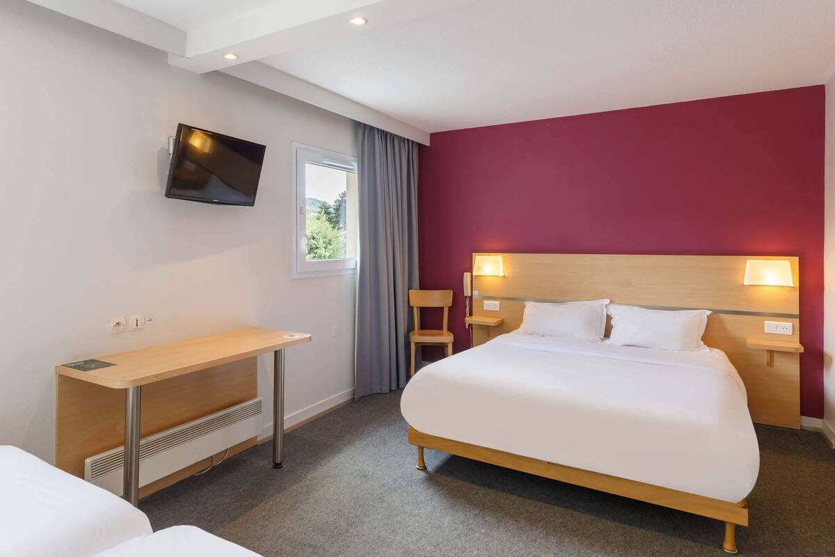 Habitació d'hotel amb llit, escriptori, televisió i finestra amb cortines.