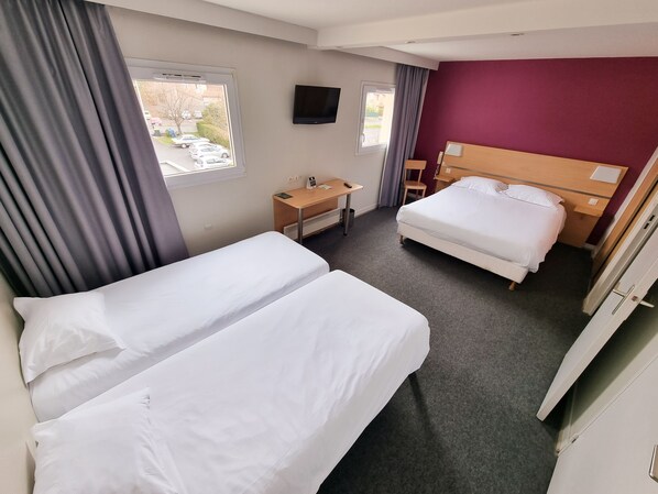 A hotel room with two beds, a desk, a TV, and a window with a view of parked cars.