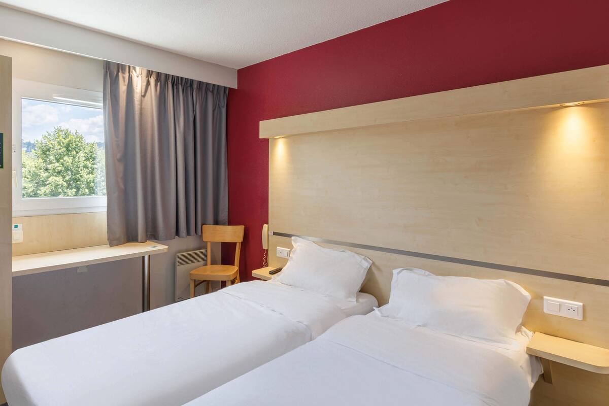 Habitació d'hotel amb dos llits, una finestra amb cortines, una taula i una cadira.