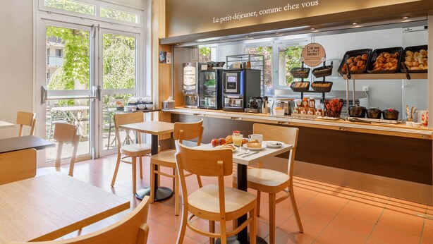A café with wooden furniture, a breakfast bar with pastries, and a view of greenery outside.