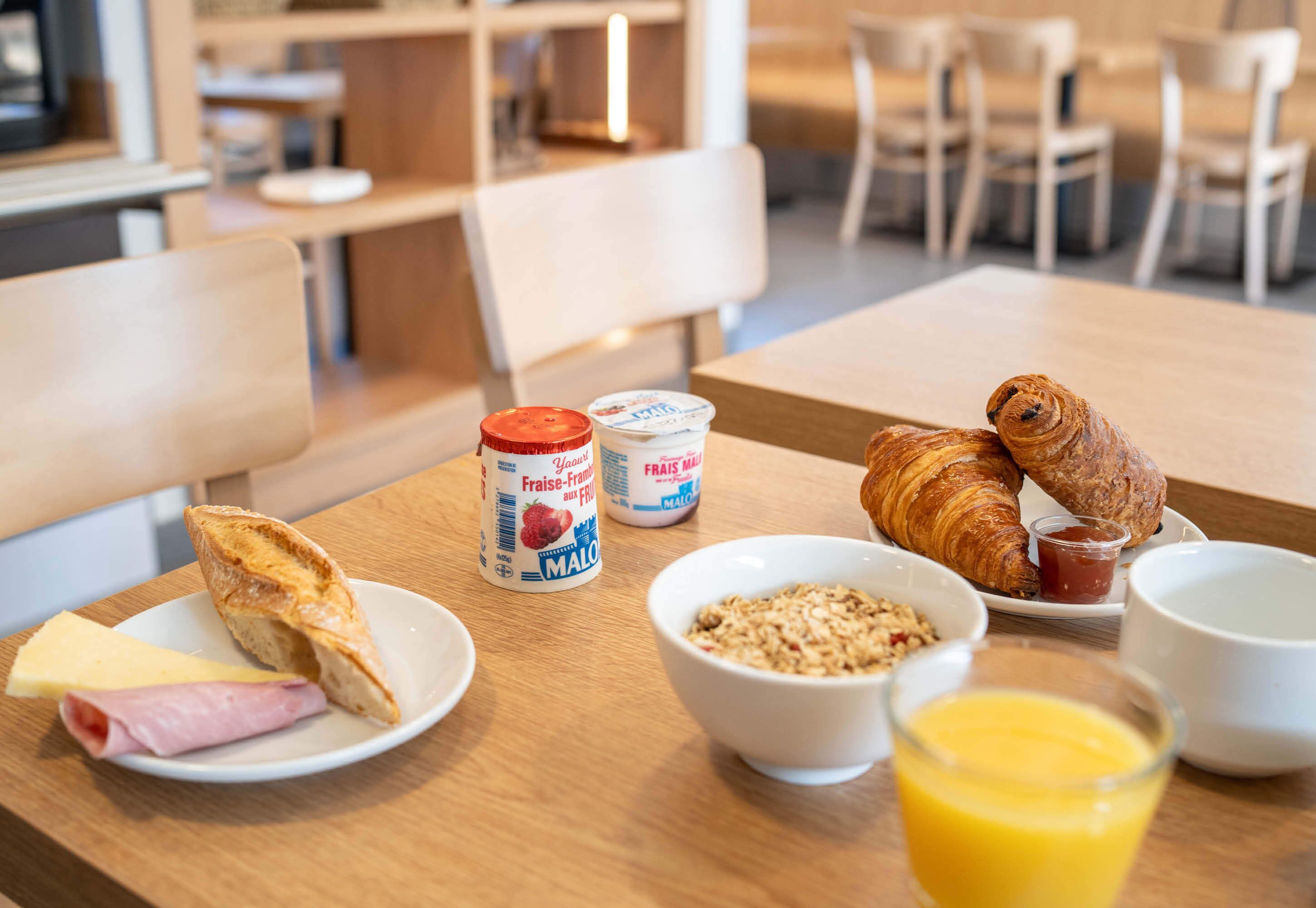 Disposição para o café da manhã com croissants, presunto, queijo, iogurte e cereal sobre uma mesa de madeira.