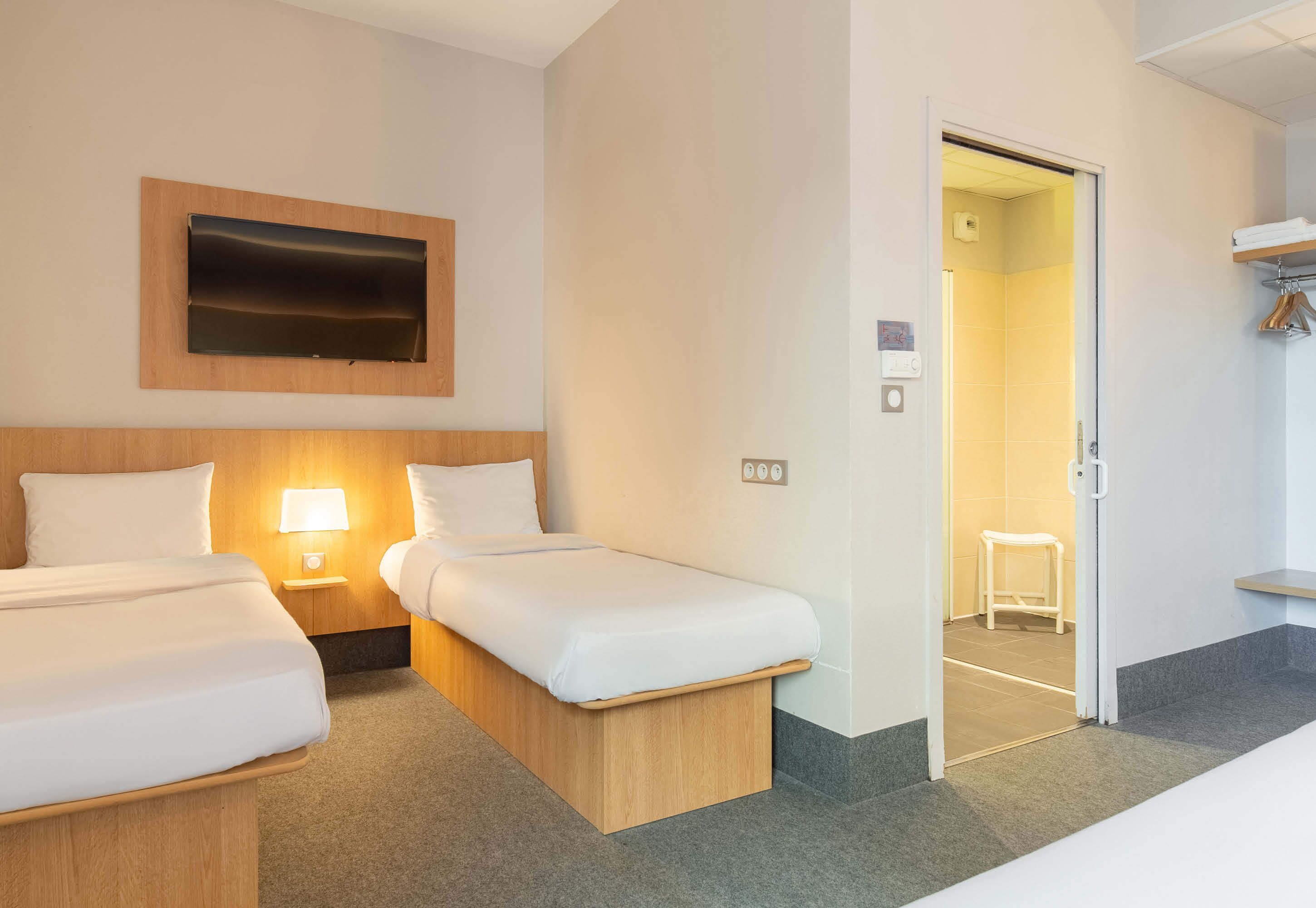 A hotel room with two beds, a TV mounted on the wall, a bedside lamp, and a small stool.