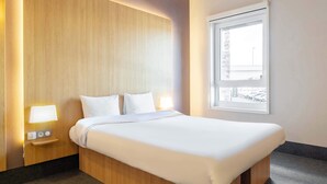 A hotel room with a wooden headboard, a large bed with white bedding, a nightstand with a lamp, and a window with a view of the city.