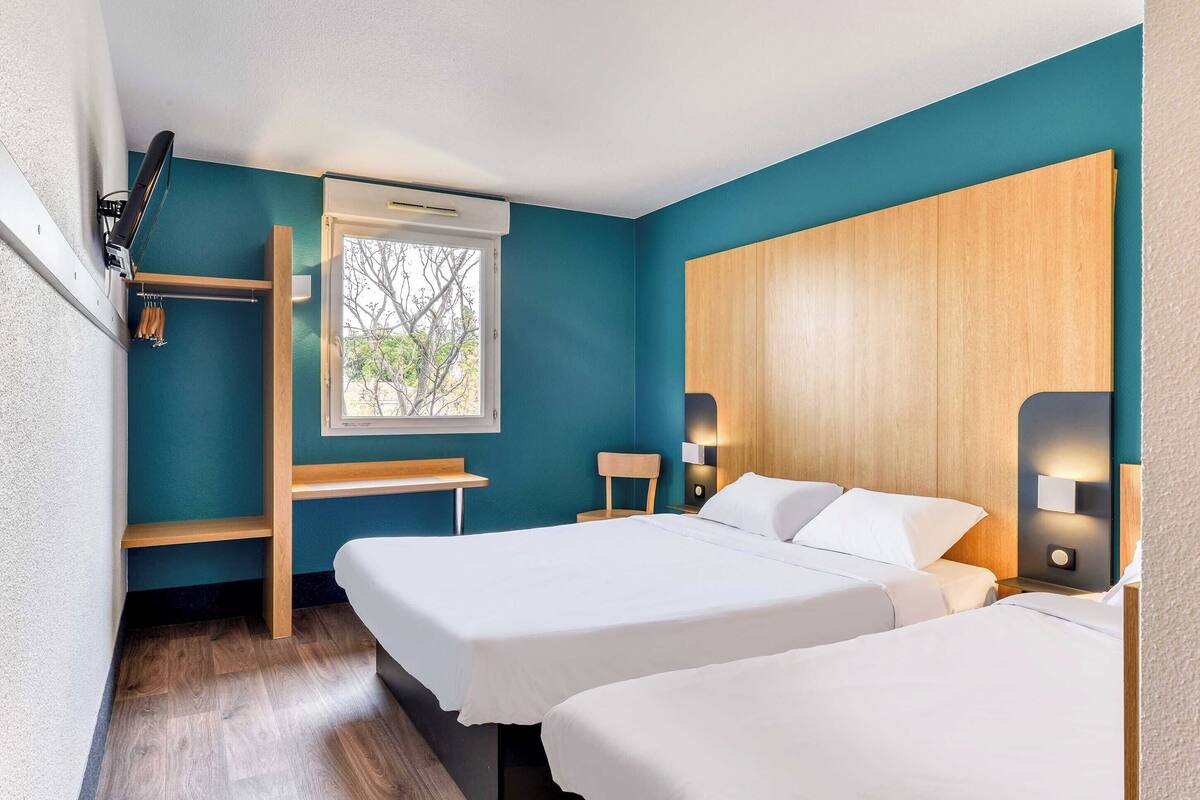 Une chambre d’hôtel aux murs de couleur turquoise, avec deux lits, un bureau en bois, une fenêtre donnant sur un paysage agréable et une étagère intégrée.