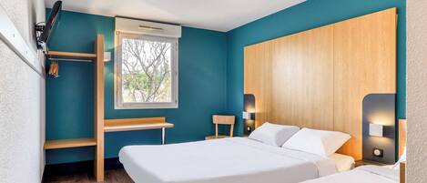 Una habitación de hotel con paredes color turquesa, dos camas, un escritorio de madera, una ventana con vistas y una estantería empotrada.