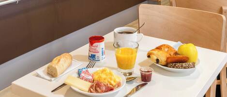 Una mesa de desayuno con pan, queso, jamón, huevos revueltos, un croissant, un limón, un vaso de zumo de naranja, una taza de café y salsas.