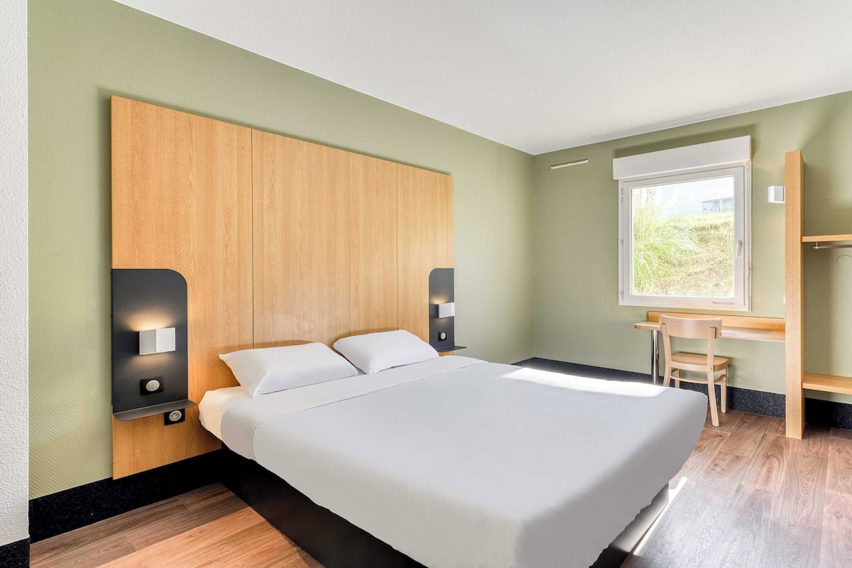 Une chambre d’hôtel moderne avec un grand lit, une tête de lit en bois, des tables de chevet avec des lampes, un bureau avec une chaise et une fenêtre donnant sur un paysage agréable.