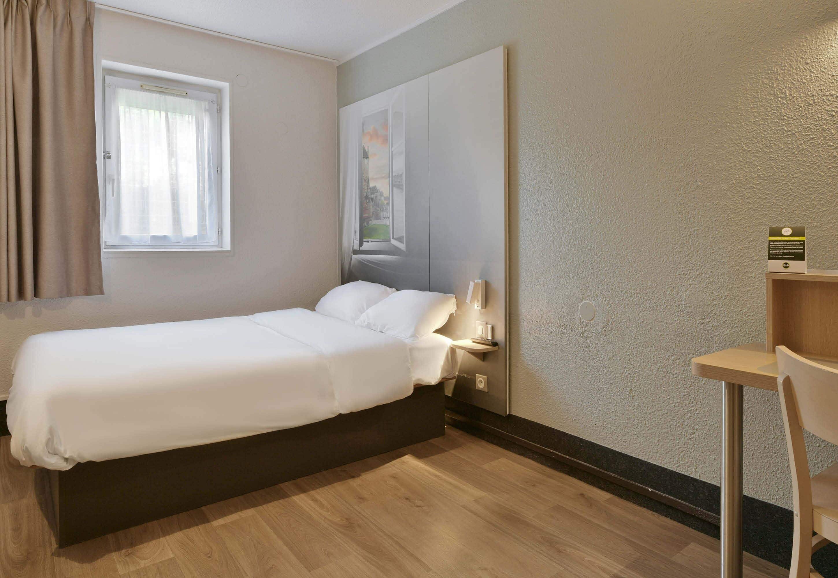 Habitación de hotel con cama, ventana con cortinas, estante en la pared y una silla.