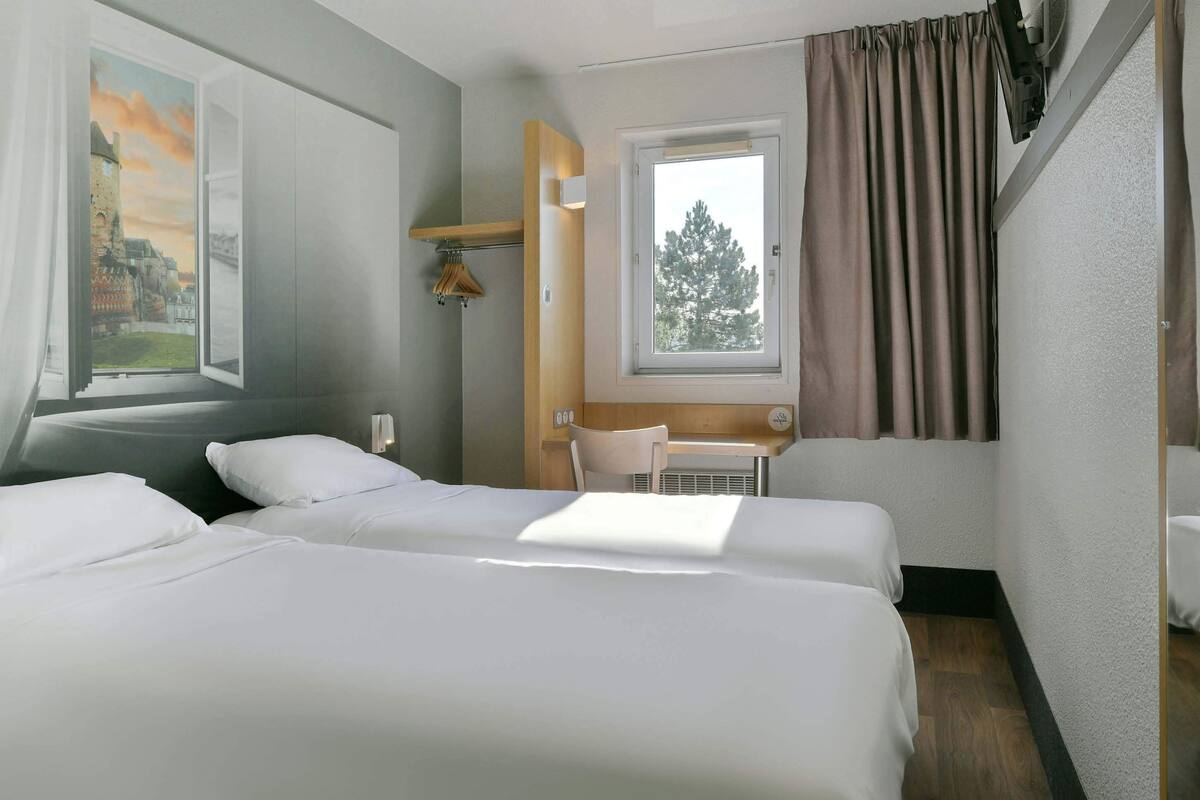 A hotel room with two beds, a window with curtains, a chair, and a small table.