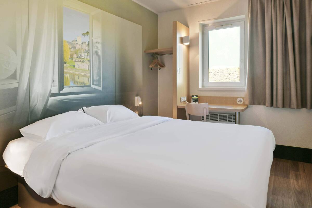 A hotel room with a large bed, a desk, a chair, a window with curtains, and a wall-mounted heater.