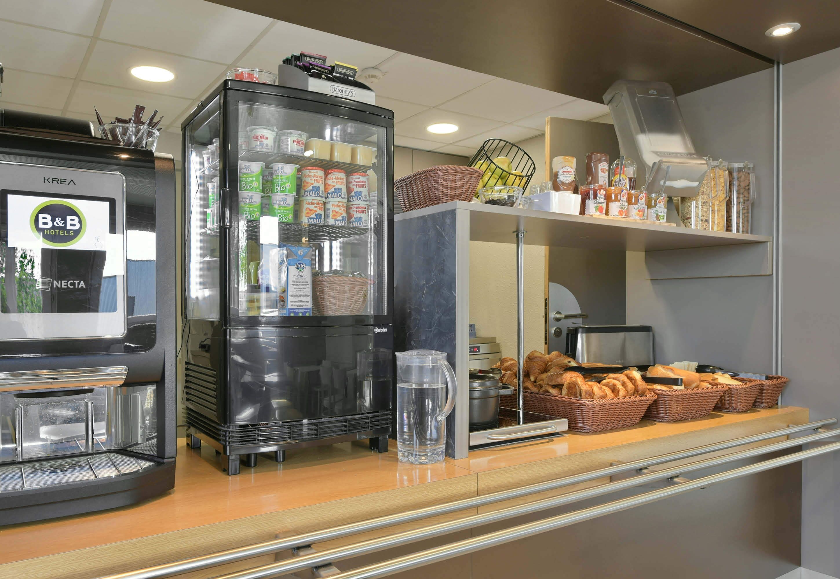 A hotel breakfast area with a coffee machine, refrigerator stocked with beverages, and a display of bread and pastries.