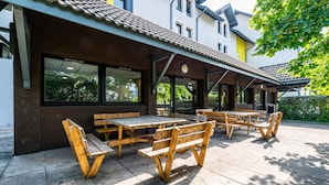 An outdoor patio with wooden tables and benches, a covered seating area, and a building with large windows and a tiled roof. - B&B HOTEL Chambéry La Cassine (Chambery)