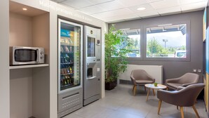 A modern waiting area with vending machines, a microwave, seating, and a view of the outdoors. - B&B HOTEL Chambéry La Cassine (Chambery)