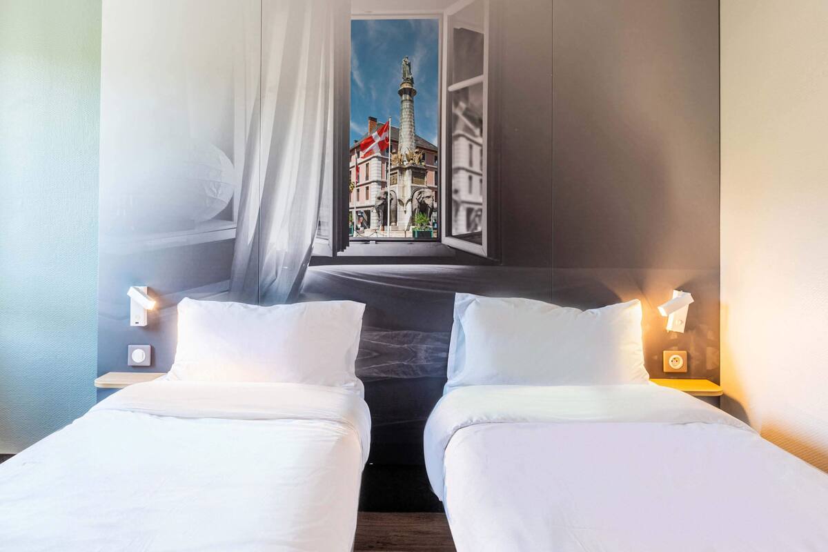 Une chambre d’hôtel avec deux lits, une fenêtre donnant sur un immeuble et des lampes de chevet.
