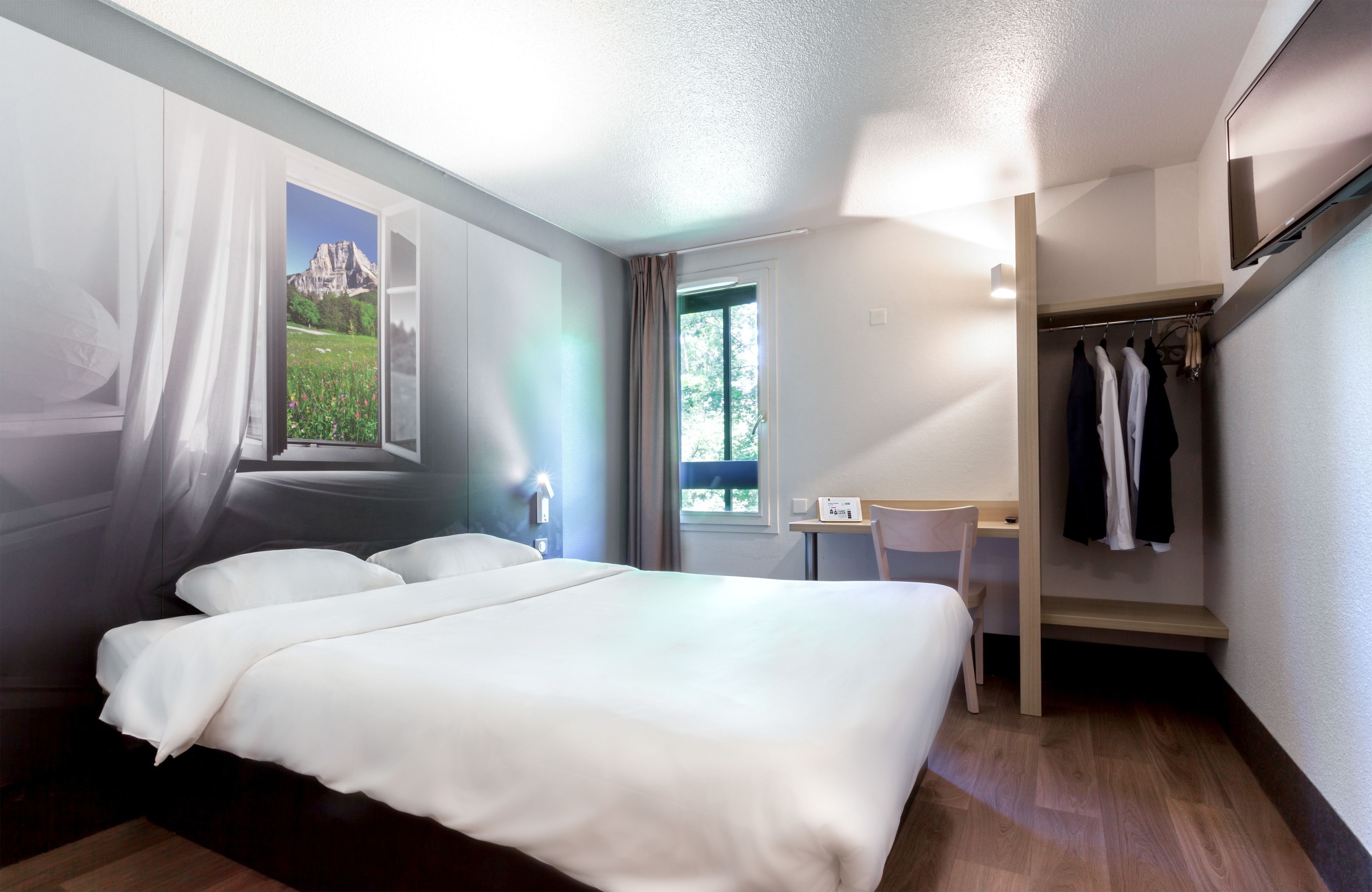 Quarto de hotel com cama, escrivaninha, cadeira, guarda-roupa e janela com vista para as montanhas.