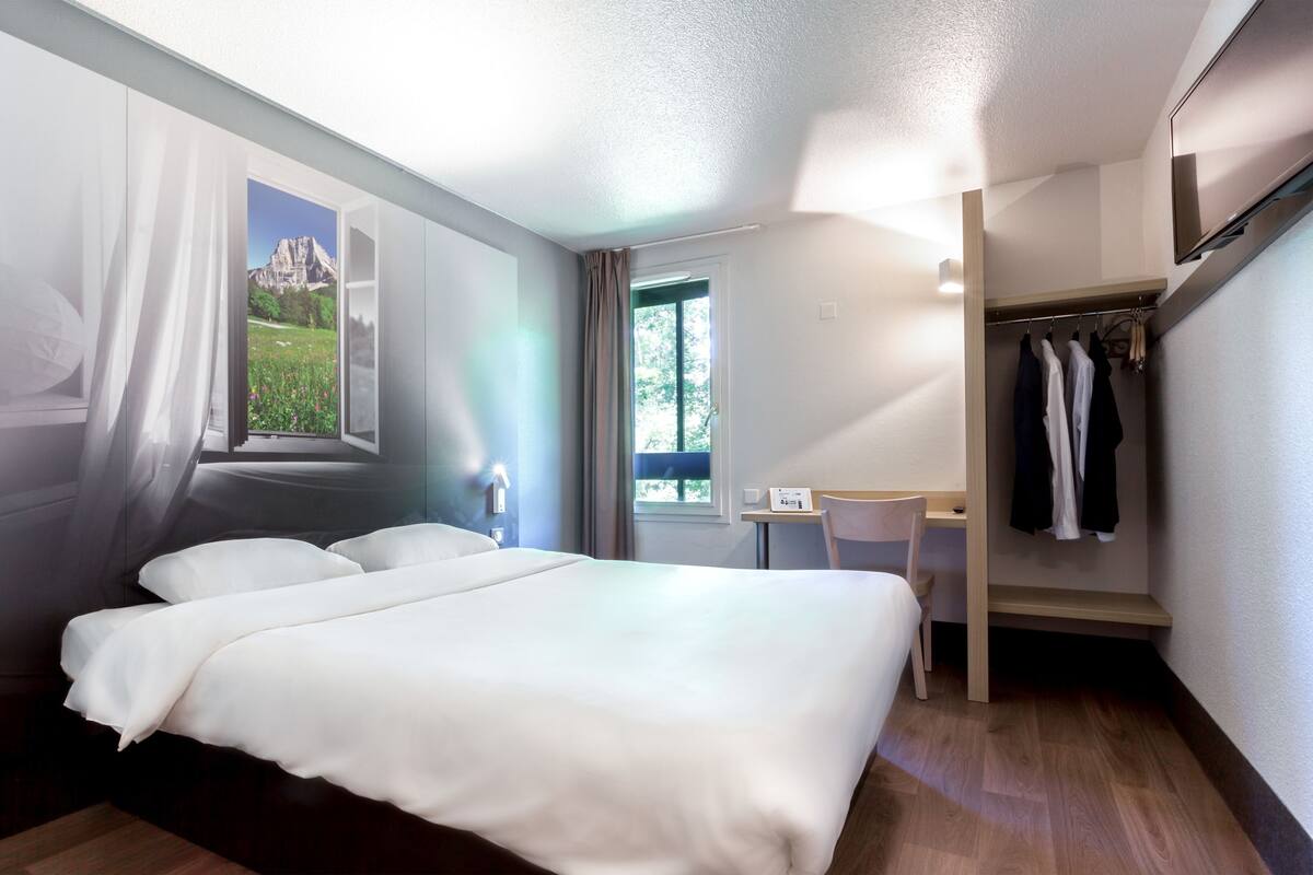 Une chambre d’hôtel équipée d’un lit, d’un bureau, d’une chaise, d’une armoire et d’une fenêtre donnant sur la montagne.