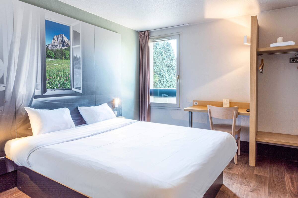 Une chambre d’hôtel avec un grand lit, un bureau et une vue sur les montagnes par une fenêtre ouverte.