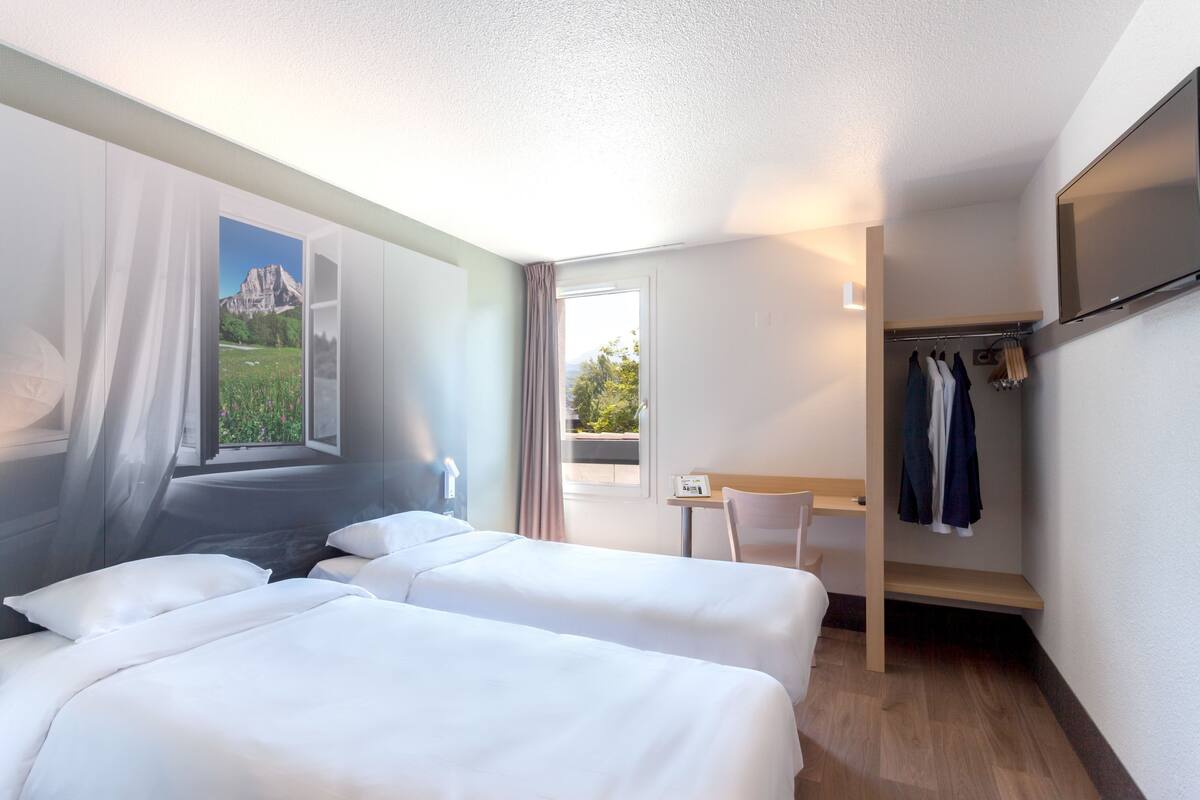 Une chambre d’hôtel avec deux lits, un bureau, une chaise, un porte-manteau et une grande fenêtre donnant sur les montagnes.