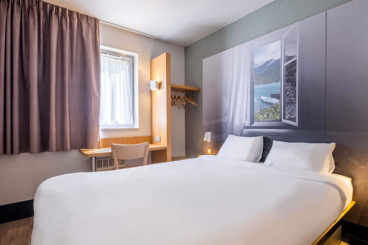 A hotel room with a large bed, a desk with a chair, a window with a view of mountains and water, and a coat rack.