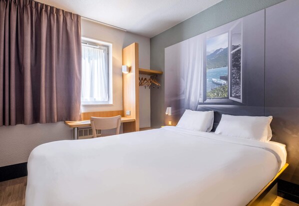 A hotel room with a large bed, a desk with a chair, a window with a view of mountains and water, and a coat rack.