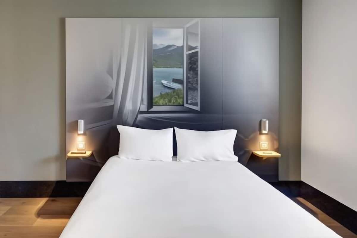 A bedroom with a bed, two bedside lamps, and a large window with a view of a lake and mountains.