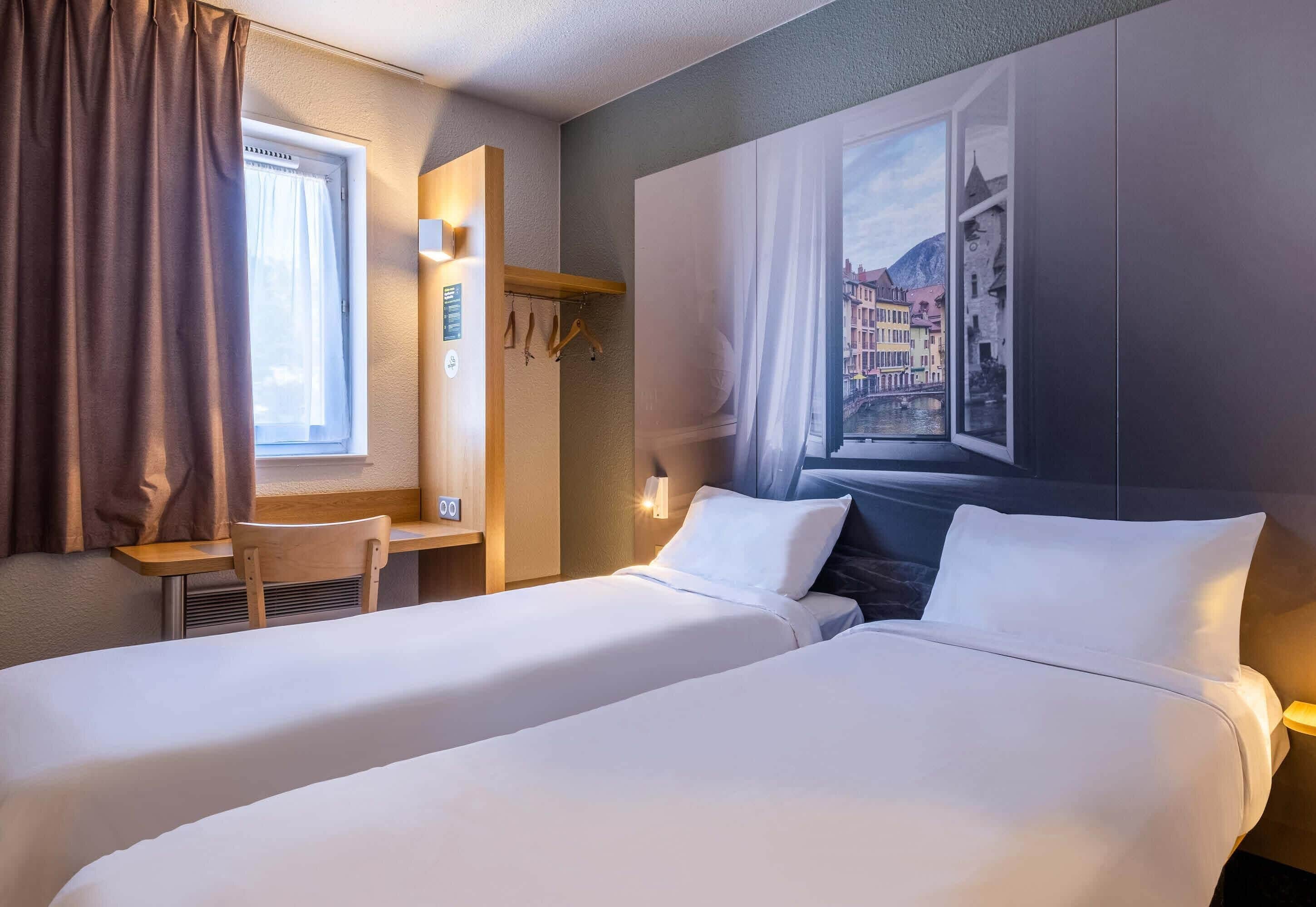 A hotel room with two beds, a desk, a chair, a window with curtains, and a wall-mounted TV.