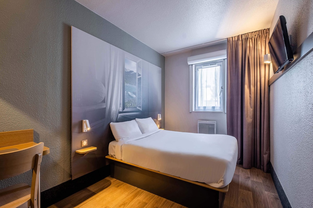 A hotel room with a bed, bedside table, lamp, and a TV mounted on the wall.