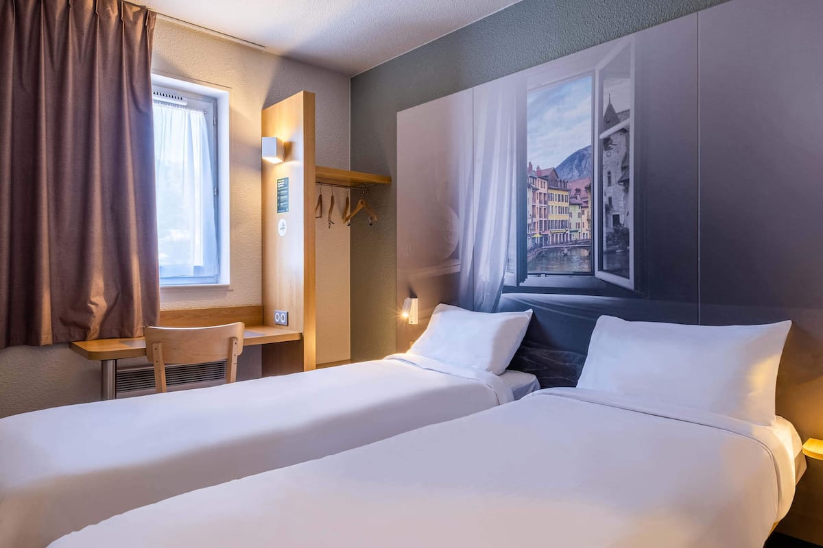 A hotel room with two beds, a desk, and a window with a view of buildings.