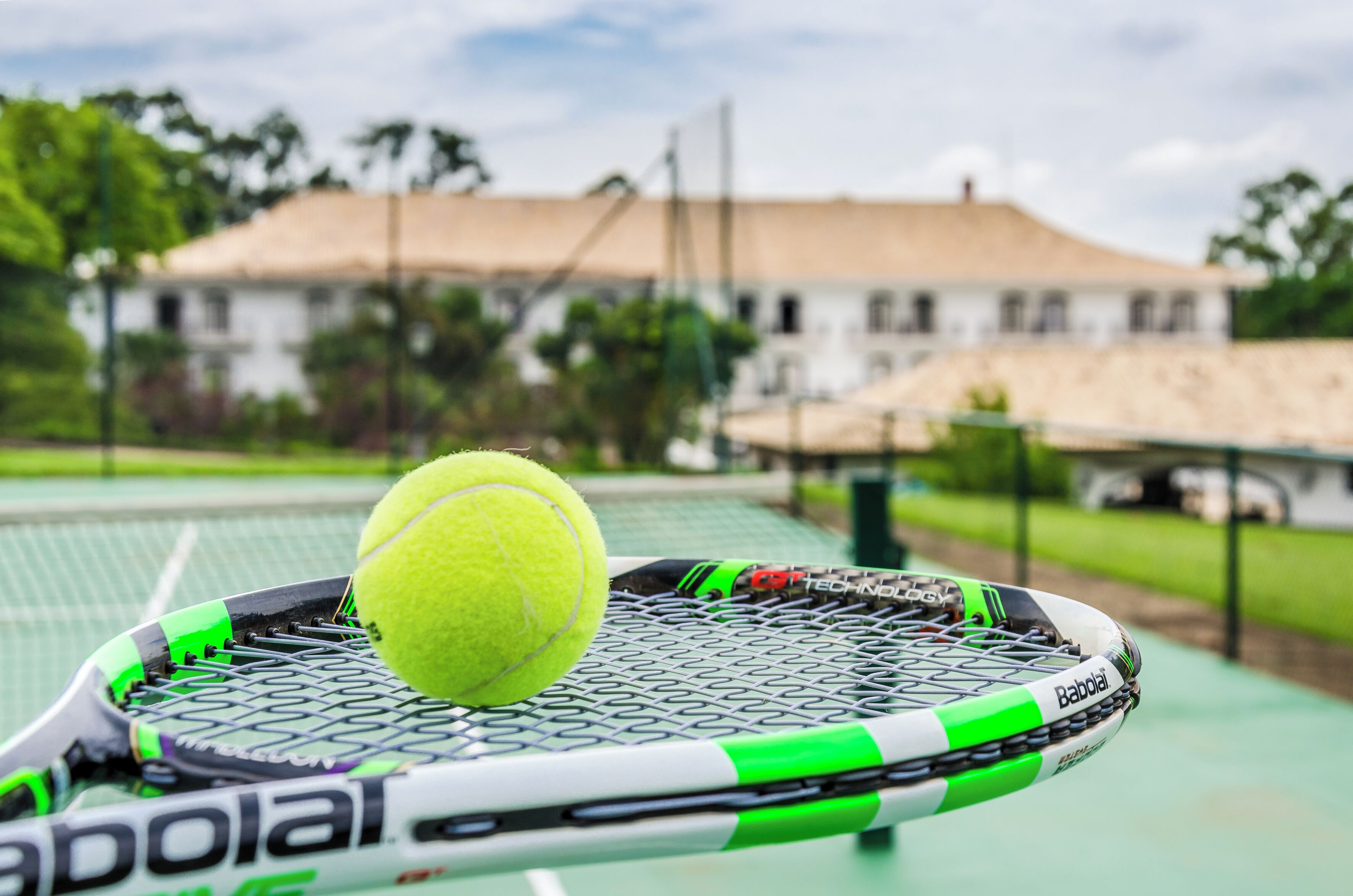A tennis racket with a ball on it, a green and white tennis court, and a building with a thatched roof in the background.
