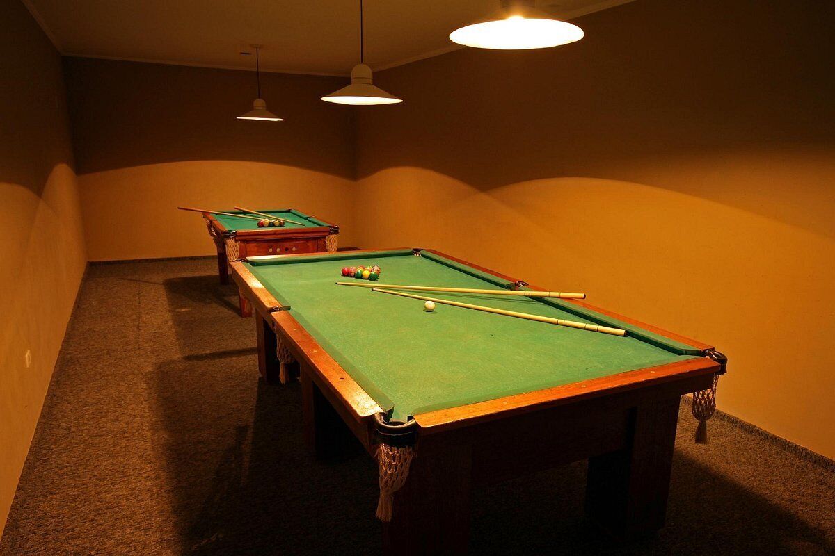 Uma mesa de bilhar com bolas e tacos, outra mesa ao fundo e piso coberto por carpete.