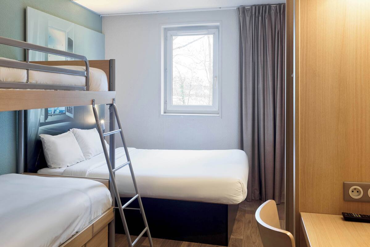 Ein kleines Hotelzimmer mit Etagenbett, Einzelbett, Schreibtisch und einem Fenster mit Vorhängen.