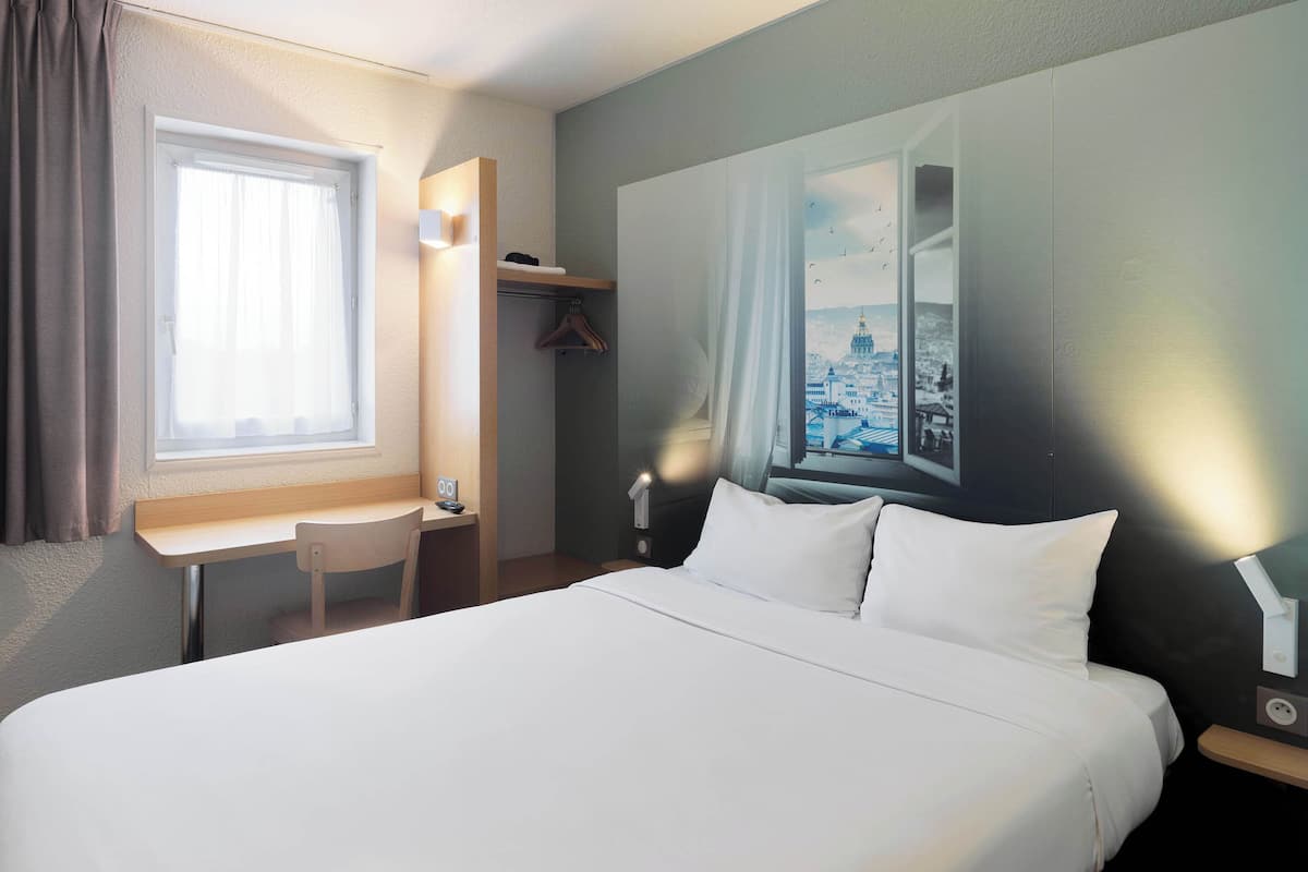 Ein Hotelzimmer mit einem Bett, einem Schreibtisch und einem Stuhl. Es gibt ein Fenster mit Vorhängen, ein Wandregal und eine Wandlampe.