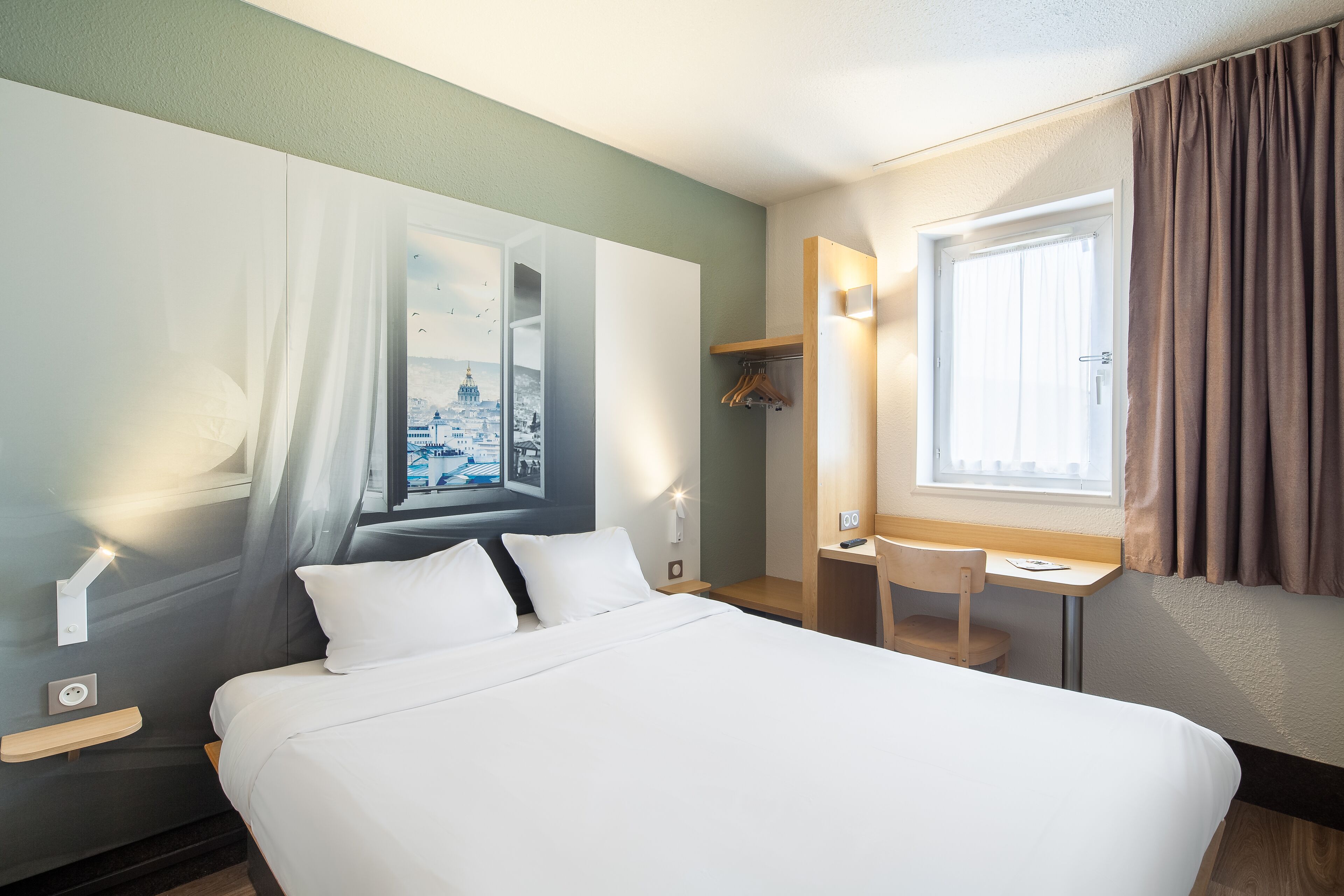 Una habitación de hotel con cama, escritorio, silla, ventana con cortinas y una lámpara de pared.