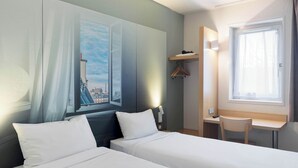 A hotel room with two beds, a desk, a chair, a window with curtains, a coat rack, and a painting of a cityscape. - B&B HOTEL Saint-Michel-sur-Orge (Saint-Michel-sur-Orge)
