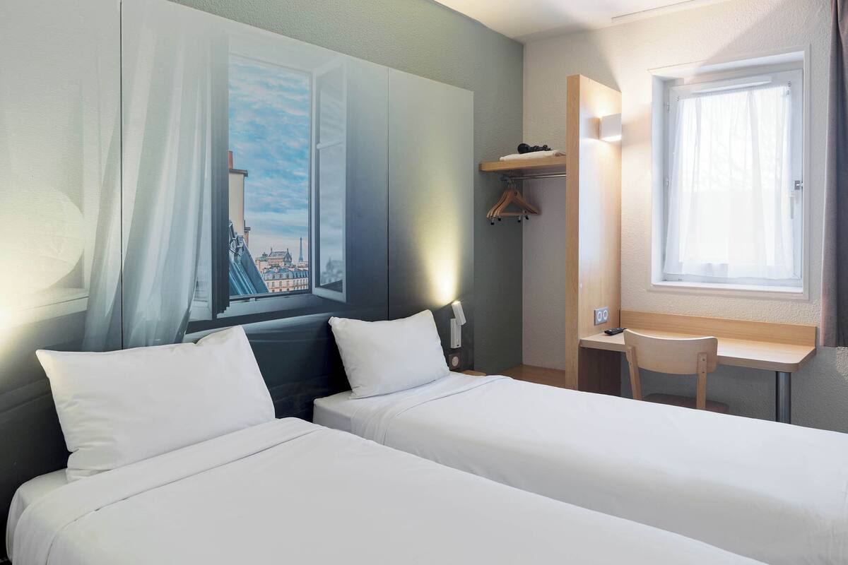 Ein Hotelzimmer mit zwei Betten, einem Schreibtisch, einem Stuhl, einem Fenster mit Vorhängen, einem Kleiderständer und einem Stadtbild.